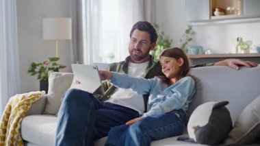 Baba kızı rahat bir kanepede oturarak internet alımları için tablet başvurusu seçiyor. Gülümseyen baba genç kız sosyal medya içerikli bilgisayarları izliyor modern daireleri birleştiriyor.