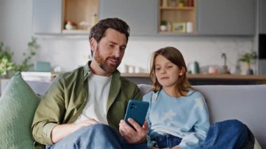 Erkek kızın uygulamalı cep telefonuna göz gezdirip daire kanepesini kapatıyordu. Gülümseyen baba, genç kıza online cep telefonu uygulamasını evde kullanmayı öğretiyor. Aile yaşamında modern teknoloji.