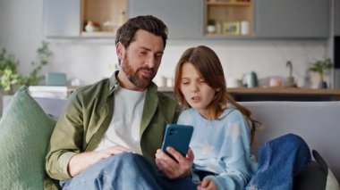 Babam akıllı telefonlu kıza oturma odasındaki rahat kanepeyi gösteriyor. Gülümseyen baba genç kızına online cep telefonu uygulaması ev koltuğu kullanmayı öğretiyor. Mutlu aile arama cep telefonu.