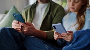 Baba çocuk sosyal medyada cep telefonlarını karıştırıyor. Apartman dairesinin iç kısımlarını kapatıyorlar. Gülümseyen adam online içerikli genç kızın rahat kanepesini gösteriyor. Modern aile akıllı telefon kullanıyor..