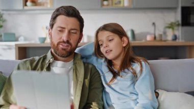Baba kızı internet üzerinden tablet başvuru kanepesini kapatarak alışveriş yapıyor. Gülümseyen baba küçük sevimli kız sosyal medya içeriğini tartışıyor rahat koltukta oturup bilgisayarını izliyor. Aile eğlencesi.