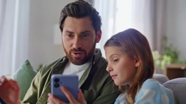Kız, babasının ekranını paylaşıyor. Rahat aile oturma odasında akıllı telefonlara bakıyor. Gülümseyen adam kızı arıyor rahat koltuklarda dinleniyor. Aile dijital online eğlence.
