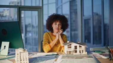 Mimari tasarımcı, modern ofiste masanda bina modeli ile poz veriyor. Gülümseyen Afro-Amerikan kadın mimar emlak ajansının işyerinde oturuyor. Kameraya başarılı bir mühendis bakımı