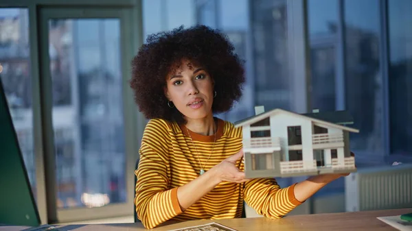 Mülk yöneticisi, modern bina modelini kapalı tutarak iş yerinde poz veriyor. Gülümseyen Afro-Amerikan mimarın portresi ev düzenini sunar. Başarılı kıvırcık kadın tasarımcı emlak ofisinde oturuyor.