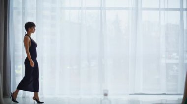 Uzun elbiseli rahat bir kadın panoramik düz oturma odasında yürüyor. Zarif kısa devre modeller şehir manzaralı pencere önünde toplanıyor. Kokteyl partisi kıyafetli kendine güvenen bir kadın dairede tek başına dolaşıyor.