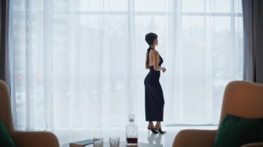 Huzurlu kadın perdelerin arasından bakıyor. Geniş, parlak yaşam alanı. Şık, kısa metrajlı, manzaralı şehir manzaralı pencere toplantısı hazır. Kesik elbiseli, kendine güveni tam bir kadın.