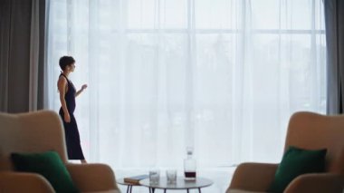 Sakin bir kadın büyük bir pencerede yürüyordu. Şık, kısa metrajlı, gökyüzüne bakan şehir manzaralı perdelere dokunan modern oturma odası. Akşamları kendine güveni olan bir bayan Dümdüz yalnız dolaşır