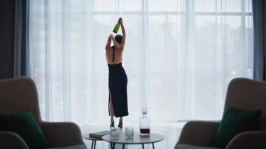 Dans eden sarhoş kadın panoramik pencere otel manzarası. Uzun ve zarif bir elbise giymiş, elinde boş bir şişe tutan yalnız bir kadın şeffaf perdelerin iç tarafında yavaşça hareket ediyor. İnce model, alkolle gevşetilmiş.