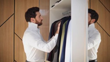 Şık bir adam iş kıyafetini seçiyor. Hafif gardırop yakın plan. Sakallı adam soyunma odasında askılara tek başına dokunuyor. Ciddi bir şekilde odaklanmış esmer beyefendi yıldönümü kutlamasını içeride yapmayı planlıyor.