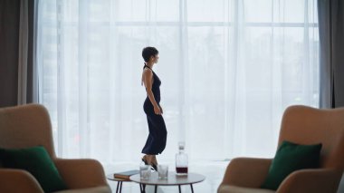 Zarif bir kadın uzun bir gecelik giyiyor panoramik düz iç mekan. Zarif kısa devre model şehir manzarası penceresinden randevu bekliyor. Tek başına kokteyl kıyafeti giyen kendine güvenen bir kadın.