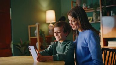 Gülümseyen anne, cep telefonuna göz gezdiriyor ve yakın çekim çekilmiş bir fotoğraf videosu izliyor. Mutlu genç anne, tapılası oğlu çevrimiçi akıllı telefon oturma masasına bakıyor. Rahatlamış kadın çocuk bağı kuruyor..