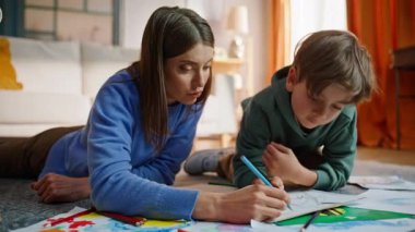 Anne, oğlunun yaratıcı aile eğlencesini çizmesine yardım et oturma odasının zeminini kapat. Özenli küçük çocuk hayal gücünü kullanarak renkli kalemlerle resim yaratıyor. Anne çocuğu sanat hobisiyle uğraşıyor..