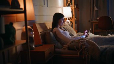 Rahat bir yatakta tablet kullanan uykusuz bir kadın. Rahat genç esmer, internet uygulaması bilgisayarlı mesajlaşma sitesi kullanarak rahat yatak odası aydınlatmalı lamba yakıyor. Gadgets bağımlılık kavramı.