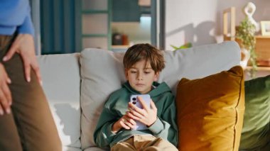 Şefkatli anne, oğlunu akıllı telefondan alıp evde oturma odasını kontrol ediyor. Üzgün küçük çocuk internetteki cep telefonu oyununa karışmış. modern anne endişeli şirin çocuk telefonu bağımlılığı.
