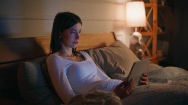 Rahat bir kadın tablet gibi görünüyor. Rahat bir daire yatağı. Yakın plan. Rahat bir uyku saatinde internetteki içerik bilgisayarını izleyen esmer bir gülümseme. Huzurlu kadın yatak odasında dijital aletlere bakıyor..