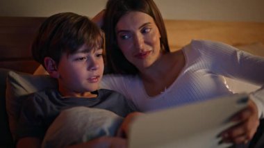 Anne oğul tablet seyrediyor, rahat bir daire yatağı, yakın plan. Gülümseyen genç anne sevimli çocuk elinde bilgisayarla akşam çizgi film seyrediyor. Mutlu annelik çocukluk konsepti.