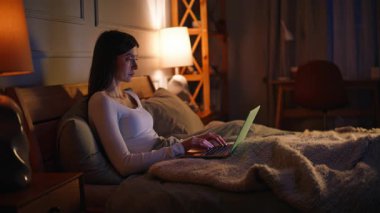 Gece çalışan ciddi bir serbest çalışan, rahat, karanlık bir yatak odasında yatıyor. Konforlu ev yatağında dizüstü bilgisayar kullanan odaklanmış genç bir kadın. Rahatlamış, çekici bir kadın çevrimiçi içerik sohbeti yapıyor..