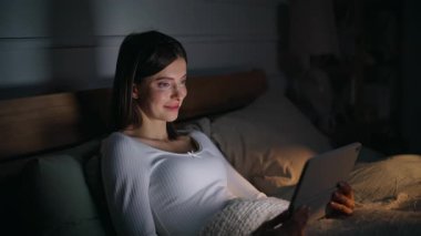 Gülümseyen esmer internette gezinme tableti içeriği rahat bir ev yatağında tek başına yatıyor. Mutlu genç bir kadın, sinema bilgisayarında sosyal medyada geziniyor, karanlık yatak odasında dinleniyor. Çevrimiçi bağımlılık.