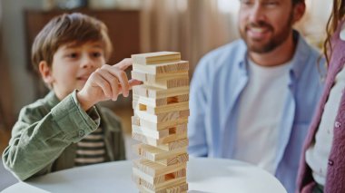 Mutlu çocuk, ailesiyle yakın planda jenga oynuyor. Gülümseyen baba flor oturan çocuğu destekliyor. Odaklanmış çocuk rahat bir hafta sonu dairesinde ahşap blokları kaldırıyor ve maçın tadını çıkarıyor. Aile aktivitesi