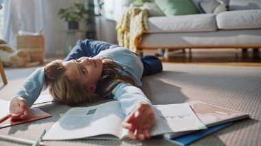Sevimli kız motivasyonsuz ev ödevi, halı sarma güneşli oturma odası, yakın çekim. Aktif liseli kız okul kitaplarının yanında yerde yatıyor. Neşeli tembel genç bayan sıkıntıdan ders çalışıyor..