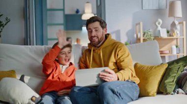 Baba oğul tableti selamlıyor. Video başlıyor. Modern ev kanepede oturuyoruz. Gülümseyen baba küçük çocuk el sallıyor bilgisayar kameraları çevrimiçi iletişimin keyfini çıkarıyor. Aile sanal toplantı sohbeti.