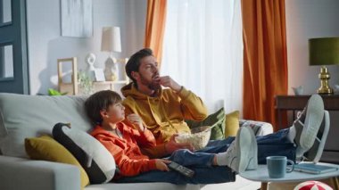 Baba çocuk komedi gülüyor rahat bir ev kanepesi. Mutlu ebeveyn çocuk televizyon izlerken apartman kanepede oturmanın tadını çıkarıyor. Patlamış mısır atan neşeli sakallı adam. Atıştırmalık yiyen sevimli bir çocuk..