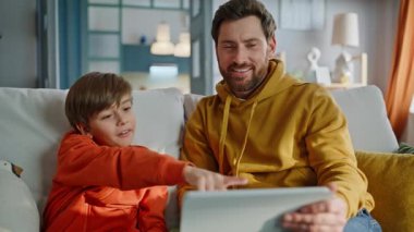 Baba çocuk tablet içeriğini tarıyor rahat kanepe kapanışı yapıyor. Gülümseyen sakallı adam, küçük bir çocukla birlikte eğlenceli bir video kaset izliyordu. Mutlu baba oğul rahatla.