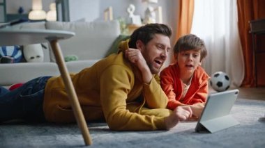 Baba çocuk çizgi film izliyor. Tablet evde yerde yatıyor. Yakın plan. Mutlu baba mutlu oğul gülerek internetteki komik bilgisayarın keyfini çıkarıyor. Duygusal ebeveyn erkek aile dijital aygıtlara göz atıyor.