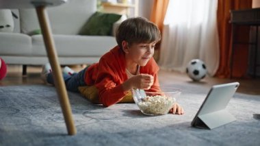 Rahat çocuk karikatür tableti izliyor oturma odasındaki halıyı tek başına yakından çekiyor. Ev işlerinde patlamış mısır yiyen şirin küçük bir çocuk. Mutlu çocuk çevrimiçi eğleniyor.