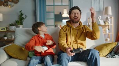 Neşeli video oyunu kazanan çocuk üzgün babasıyla oynuyor oturma odasındaki kanepeye yakın çekim yapıyor. Heyecanlı rekabetçi çocuk hayal kırıklığını hisseden babasından online zafer gülüşünü kutluyor. Aile oyunu hobisi.