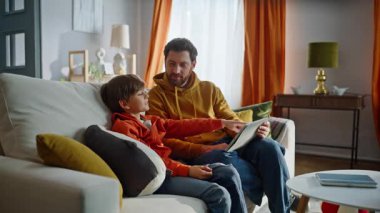 Baba oğul, oturma odasındaki kanepede oturup tablet içeriğini izliyor. Gülümseyen sakallı, sevimli, küçük bir çocuk internet alışverişi yapıyor. Ev kanepesinde bilgisayar uygulaması kullanıyor. Aile teknolojisi kullanımı
