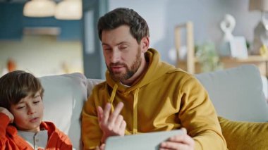 Baba oğul tablet içeriğini tartışıyor rahat bir daire kanepesi kapanışı. Gülümseyen genç baba mutlu oğul internetten alışveriş yapıyor bilgisayarın e-ticaret uygulamasını izliyor. Aile gereçlerle evi rahatlatıyor.