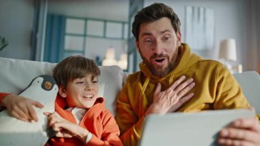 Baba çocuk oturma odasındaki kanepeye yakın otururken video tableti izliyor. Mutlu baba gülümseyen küçük oğul çevrimiçi içeriğin tadını çıkarıyor. Sakallı yakışıklı çocuk bilgisayar içeriğine duygusal tepki veriyor..