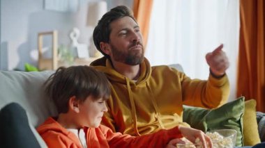 Baba çocuk komedi seyrediyor. Kaygısız aile istirahatinin keyfini çıkarıyor. Rahat kanepe kapanışı. Gülümseyen sakallı baba patlamış mısır fırlatıyor. Sevimli küçük çocuk birlikte televizyon filminden gülüyor. Mutlu çocukluk.