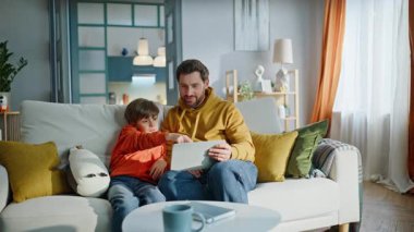 Erkek evlat oturma odasındaki kanepede oturup video tableti izliyor. Mutlu sakallı baba çocuk rahat bir evde eğitici online içeriği tartışıyor. Gülümseyen baba çocuklara teknoloji kullanmayı öğretiyor..