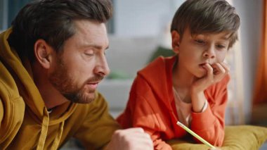 Erkek öğrenci ders kitabı okuyor. Oturma odası zeminine yakın çekim yapıyor. Sakallı babayla okul dersini öğrenen tatlı bir çocuk. Gülümseyen baba oğlanın kafasını okşuyor..