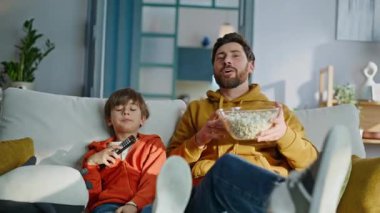 Sevimli oğlunun uzaktan kumandayla televizyonu açması sevimli sakallı babasının yakın planıyla rahat koltukta oturması. TV izleyen ebeveyn çocuk patlamış mısır yiyerek eğleniyor. Tasasız aile eğlencesi..