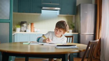 Mutfak masasında oturan düşünceli öğrenci konuyu öğreniyor. Akıllı ilkokul öğrencisi ders yazmayı düşünüyor. Ev ödevi yapıyor. Şirin küçük çocuk içeride ödev çalışıyor.