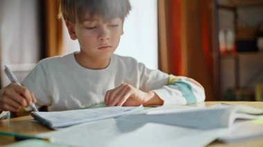 School student writing pen making notebook notes sitting kitchen table closeup. Diligent elementary pupil learning homework reading subject material home. Intelligent little boy studying lesson alone.