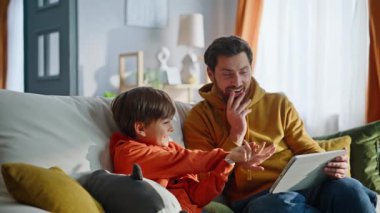 Aile, evde rahat rahat oturup komik video tabletleri izliyor. Gülümseyen ebeveyn çocuk çevrimiçi içerik görünümlü cihaz ekranını birlikte seviyor. Mutlu baba, sevimli çocuk bilgisayarını karıştırıyor, kanepeyi rahatlatıyor..