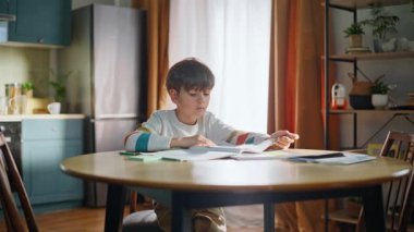 Smart child learning lesson doing homework at modern home kitchen closeup. Intelligent schoolboy writing notebook flipping pages thinking school task sitting table. Children home education concept.
