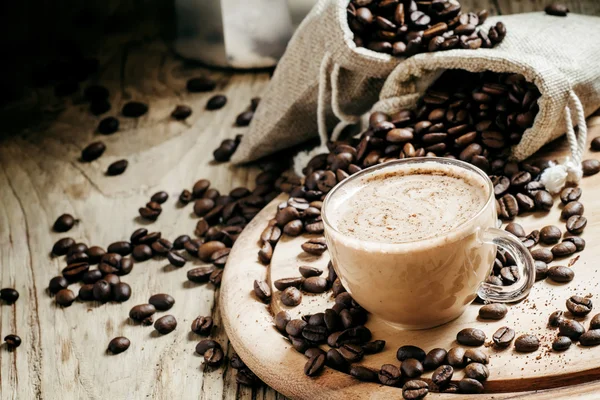 Café con leche en una taza de vidrio, granos de café en bolsas de lona —  Fotos de Stock