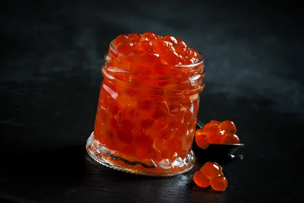 Caviar vermelho em um frasco de vidro no fundo de pedra preta — Fotografia de Stock