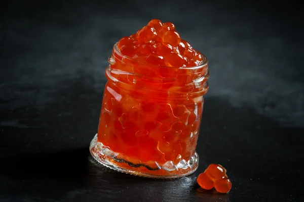 Caviar vermelho em um frasco de vidro no fundo de pedra preta — Fotografia de Stock