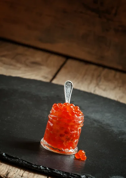 Caviar vermelho em um frasco de vidro no fundo de pedra preta — Fotografia de Stock