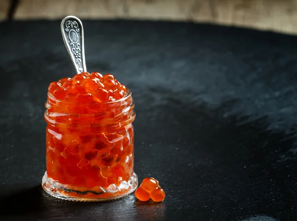 Caviar vermelho em um frasco de vidro no fundo de pedra preta — Fotografia de Stock