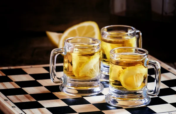 Whisky with lemon in old-fashioned glass mugs on a chess board — Stock Fotó