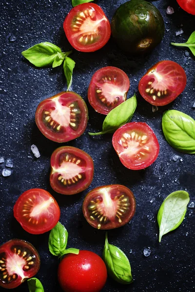 Mitades rojas y negras de tomates con hojas verdes de albahaca — Foto de Stock