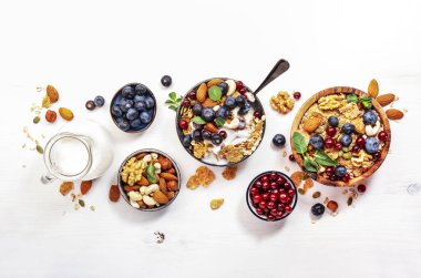 Müsli kasesi ve sağlıklı kahvaltı için organik malzemeler. Granola, fındık, yabanmersini, yaban mersini, yulaf ezmesi, Yunan yoğurdu, beyaz masada tam tahıl gevreği.