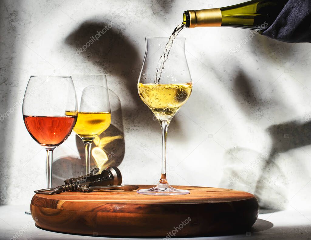Wine tasting. White wine pouring into glass on background with selection of red, white and rose wines in glasses. Hard sunlight and shadows from foliage